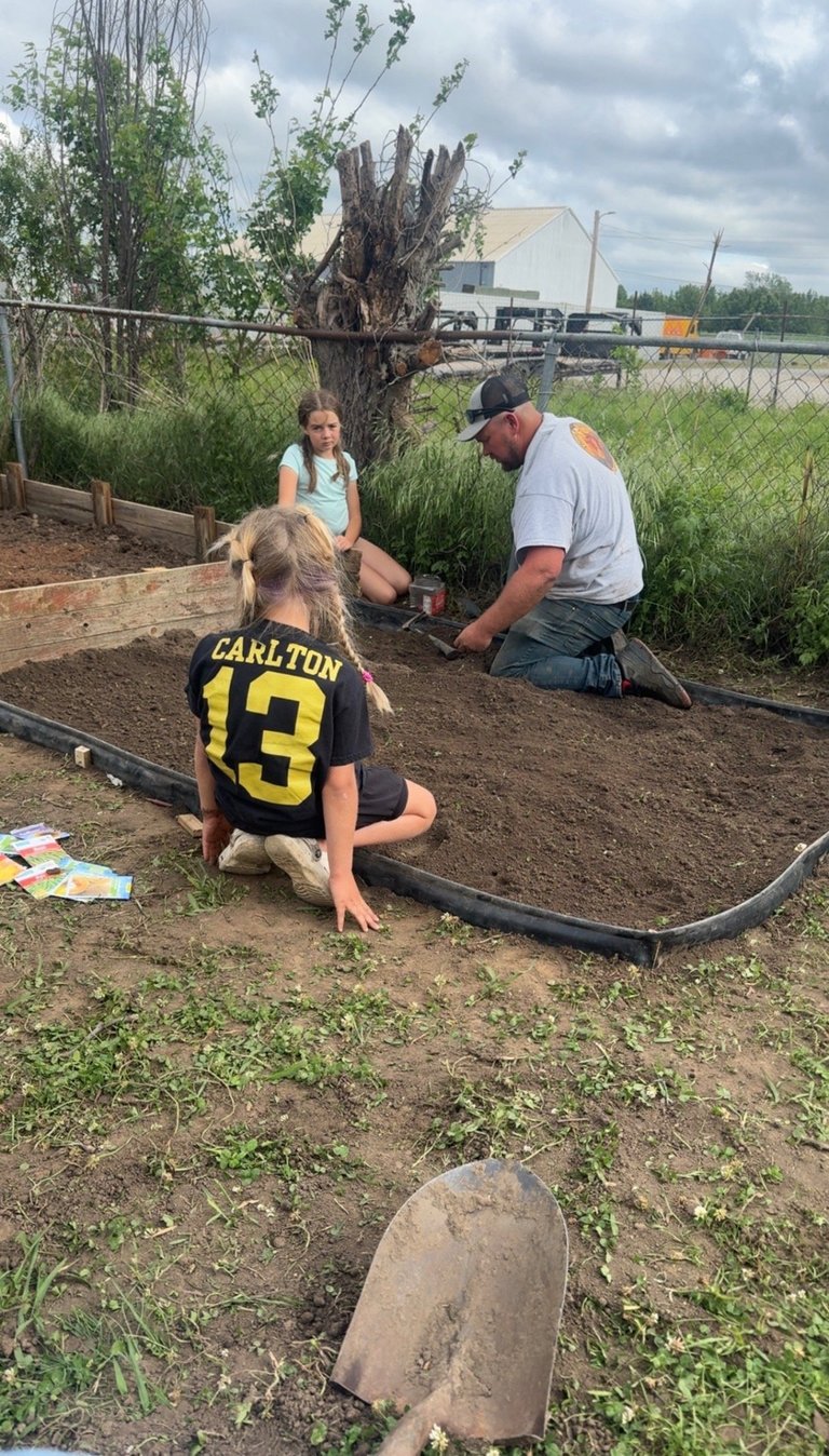 An adult and children working in a garden, kneeling by a tree and raised bed with gardening tools nearby