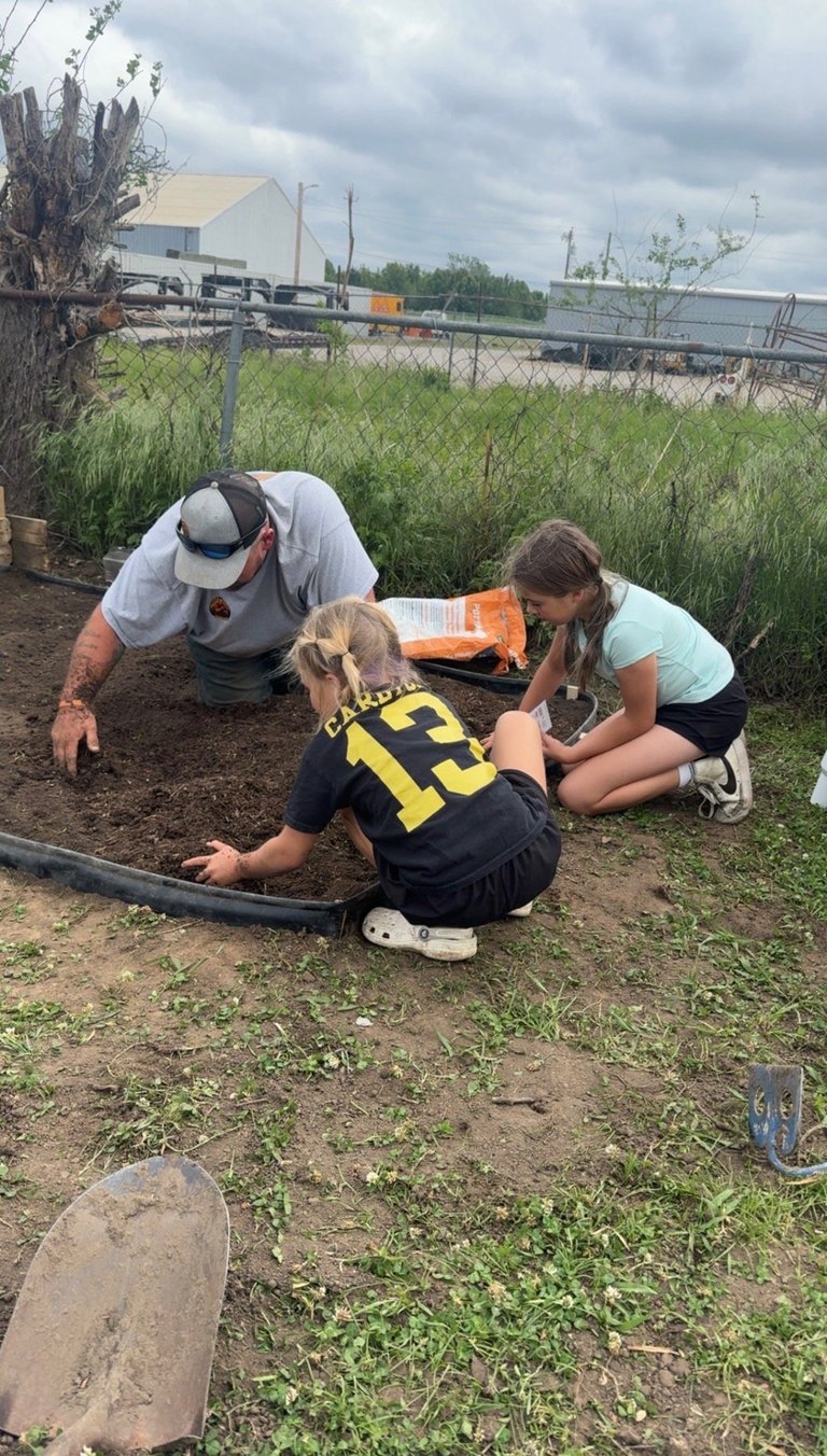 Two children and an adult examining something on the ground in a garden area with a fence and industrial buildings in the background