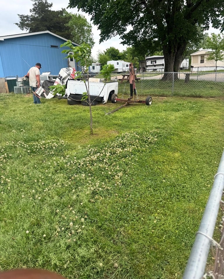 People planting a young tree in a grassy yard with a white trailer, blue shed, and mobile homes in the background
